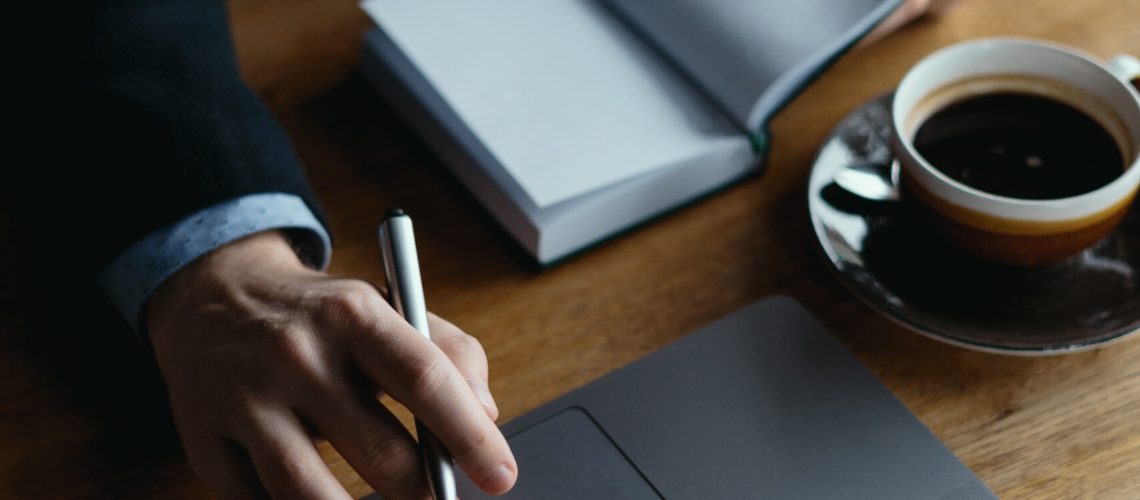 Business man hand on a laptop touchpad holding a pen and a textbook for notes researching for a project